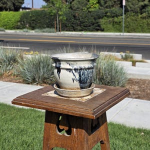May include: A ceramic planter with a matching saucer sits on a wooden plant stand. The planter is white with dark blue drips. The stand is made of dark wood and has a square top with decorative tiles.