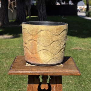 May include: A ceramic planter with a textured, wave-like pattern in shades of yellow and brown. The interior is black. It sits on a wooden stand with a decorative tile insert, against a backdrop of green grass and trees.
