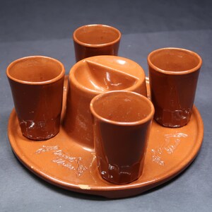 May include: A set of four brown ceramic shot glasses with a central brown ceramic holder with a small dip for a snack. The set is on a brown ceramic tray with a decorative design.