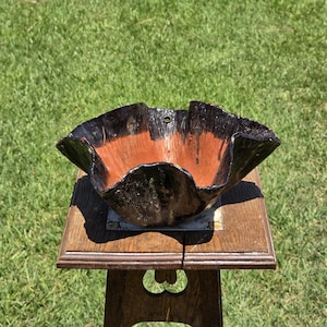 May include: A handcrafted, decorative bowl with a unique, ruffled edge design. The bowl features a striking contrast of black and reddish-orange hues. It is displayed on a small, wooden table, set against a backdrop of green grass.