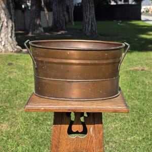 May include: A vintage copper bucket with two handles, resting on a wooden stand. The bucket has a cylindrical shape with horizontal bands and a weathered patina. The stand is made of wood and has a decorative cutout design.