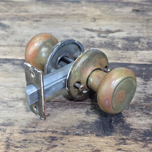 May include: A vintage door knob set featuring two tarnished brass spherical knobs connected by a square metal spindle. The assembly includes a rectangular brass faceplate with two screw holes and a protruding latch mechanism, showing signs of age.
