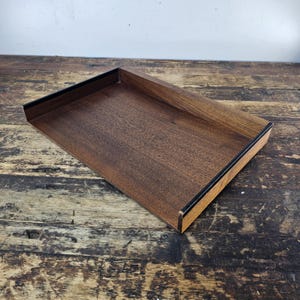May include: A rectangular wooden tray with a dark brown finish and black trim. The tray has a shallow depth and is designed with a simple, modern aesthetic. The wood grain is visible, adding texture to the surface.