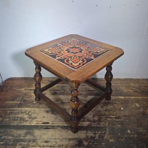 May include: A square wooden side table with a tiled top featuring an orange, black, and cream floral design. The table has turned legs and measures approximately 24 inches wide and 20 inches tall.