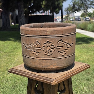 Puede incluir: Una maceta cilíndrica de terracota marrón con un motivo floral tallado en el exterior. El diseño consiste en una flor central rodeada de patrones de hojas simétricos, enmarcada por dos bandas horizontales que rodean el cuerpo de la maceta.