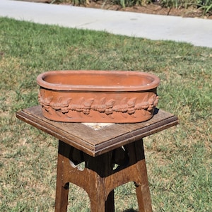 May include: An oval terracotta planter featuring a raised relief pattern of leaves and berries encircling the exterior. The planter has a smooth, flat rim and a deep, open interior, showcasing the natural reddish-brown clay texture throughout.