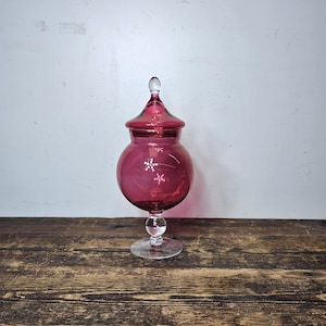 Puede incluir: Un tarro de vidrio vintage con tapa, de color rosa y base de vidrio transparente. El tarro tiene un diseño decorativo en el lateral.