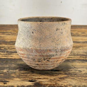 May include: A rustic, handmade pottery bowl with a wide opening and a rounded base. The bowl features a textured, light brown exterior with a band of reddish-brown stripes around the middle. The interior is a darker shade of brown.