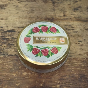 May include: A round gold-colored metal tin of Raspberry Drops with a white lid featuring red raspberry illustrations and green leaves. The text reads Raspberry Drops, Net Weight 2 1/4 ozs, Made in Israel by the Nazareth Candy Co.
