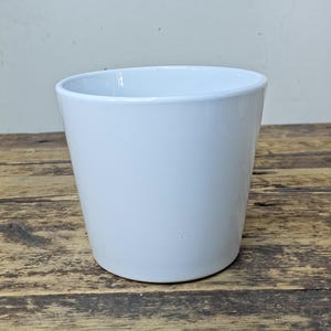May include: A white ceramic plant pot with a gently sloping form. The pot has a smooth, glossy finish and a simple, modern design. It is on a wooden surface.
