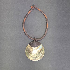 May include: A necklace featuring a large, iridescent, circular shell pendant with a dark brown cord wrapping at the top. The necklace cord is made of brown material with dark brown thread accents wrapped at intervals and a small brown bead closure.
