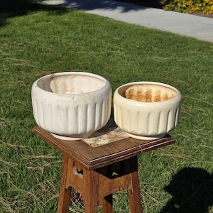 Pode incluir: Duas floreiras de cerâmica branco-sujo com detalhes canelados verticais. Uma floreira tem uma rachadura. As floreiras estão sobre uma pequena mesa de madeira com um tampo de azulejos decorativos, em um fundo de grama verde e luz solar.