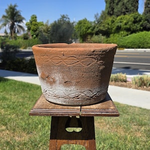 Puede incluir: Una maceta redonda de terracota con un acabado desgastado de color marrón rojizo y depósitos minerales blancos. El exterior presenta dos bandas horizontales de patrones geométricos en relieve, incluyendo semicírculos y líneas onduladas entrelazadas.
