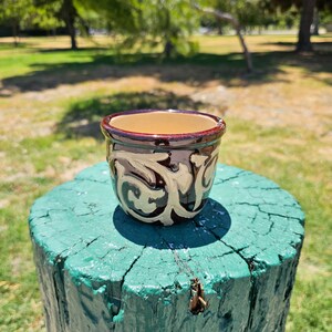 May include: A small, brown ceramic planter with a white floral design. The planter is sitting on a green tree stump.