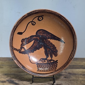 Peut inclure: Un bol rond en terre cuite avec une finition brillante, présentant une illustration peinte à la main en brun foncé représentant un oiseau perché sur un panier. Le motif inclut une ligne en spirale au-dessus de l'oiseau et une bordure décorative.