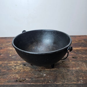Puede incluir: Un caldero de hierro fundido negro con tres patas y dos asas. El caldero está sobre una superficie de madera.