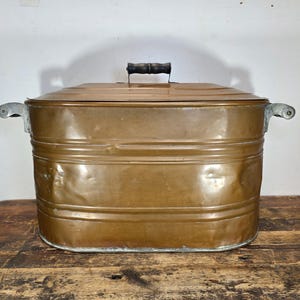 May include: A vintage copper wash tub with a lid and a wooden handle. The tub has two side handles and is rectangular in shape. The copper has a warm, aged patina, suggesting its antique nature. The tub is sitting on a wooden surface.