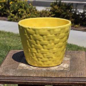 May include: A bright yellow ceramic plant pot featuring a raised, textured basket-weave pattern across its exterior surface. The vessel has a wide, circular opening and a smooth, glossy finish that reflects light, with a consistent color throughout.