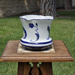 Puede incluir: Una maceta de cerámica blanca con diseños florales azules y un borde ondulado. La maceta se encuentra sobre un soporte de madera con una base de azulejos decorativos. El diseño presenta flores y hojas azules.