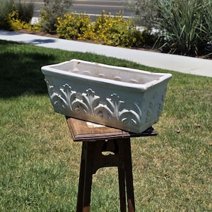 May include: A rectangular white ceramic planter featuring a raised relief pattern of stylized leaves along the exterior sides. The planter has a wide, open top and shows signs of wear with small chips in the finish along the upper rim.