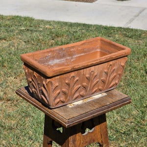 May include: A rectangular terracotta planter featuring a raised relief pattern of stylized leaves along its exterior sides. The planter has a wide, flat rim and a deep, open interior, showcasing the natural reddish-brown clay texture and finish.