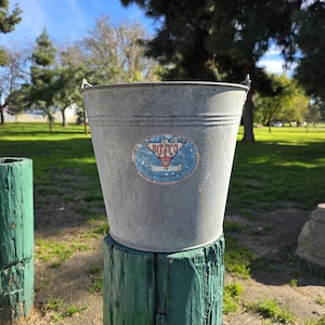 Peut inclure: Un seau en métal galvanisé avec une étiquette bleue et rouge délavée. Le seau est cylindrique avec un dessus évasé et une poignée en fil de fer. Il repose sur un poteau en bois vert vieilli dans un cadre de parc.