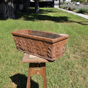 May include: A rectangular terracotta planter featuring a repeating embossed leaf pattern along its exterior sides. The planter has a wide, open top and a weathered, earthy brown finish, showcasing a textured surface with visible signs of age.