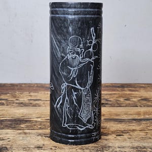 Puede incluir: Jarrón cilíndrico de mármol negro con un diseño blanco grabado de un anciano sosteniendo una rama. El jarrón tiene una superficie texturizada y está sobre una mesa de madera. El diseño es detallado y el jarrón mide unos 20 cm de alto.