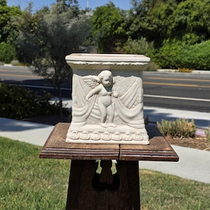 May include: A rectangular off-white ceramic planter featuring a relief sculpture of a winged cherub standing amidst draped fabric. The base and top edges are decorated with a repeating leaf pattern, and the surface has a matte finish.