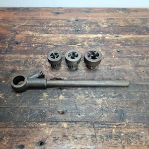 May include: A set of three vintage pipe dies with a long, metal pipe wrench. The dies are round and have a threaded hole in the center. The wrench has a handle and a long, straight shaft.