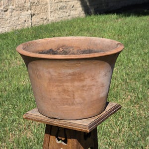 May include: A large, round terracotta planter with a wide, flared rim and a weathered, earthy brown surface. The pot features a smooth, tapered body that narrows toward the base, showing natural color variations and a dark, textured interior.