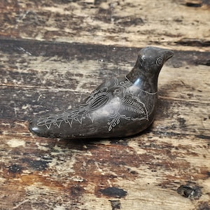 Puede incluir: Una escultura de pájaro de piedra tallada de color gris oscuro con detalles blancos grabados. El pájaro está representado en posición de descanso, con intrincados patrones en sus alas y cuerpo. La escultura está sobre un fondo de madera rústica.