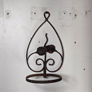 Könnte beinhalten: Ein rostiger Pflanzenhalter aus Metall mit einem dekorativen Blattdesign. Der Halter hat eine runde Basis und eine tropfenförmige Oberseite.