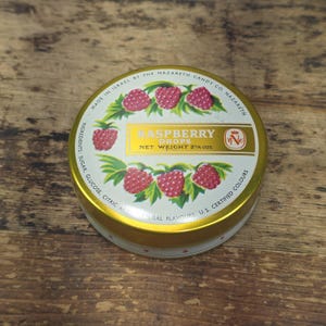 Puede incluir: Una lata redonda de metal para caramelos de frambuesa con tapa dorada. La etiqueta presenta ilustraciones de frambuesas rojas, hojas verdes y el texto Made in Israel by the Nazareth Candy Co., Raspberry Drops y Net Weight 2 1/4 ozs.