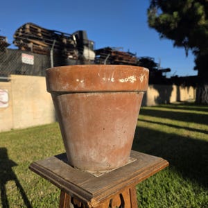 May include: A terracotta plant pot with a wide, flared rim and a tapered body. The surface of the pot displays a weathered, earthy orange-brown color with visible signs of wear, including light-colored mineral deposits and small surface scuffs.