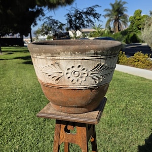 May include: A round terracotta planter featuring a wide, off-white band around the upper section with a raised, embossed floral and leaf pattern. The lower portion of the pot is a natural, unglazed reddish-brown clay with a weathered texture.