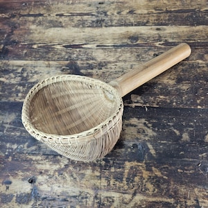 May include: A woven bamboo strainer with a long wooden handle. The strainer is brown and has a round, bowl-shaped base. The handle is attached to the side of the bowl.