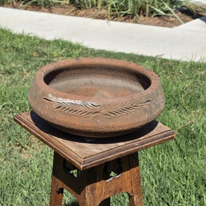 May include: A round, brown, unglazed clay bowl with a wide, thick rim. The exterior of the bowl features a carved, horizontal leaf-like pattern encircling the side. The bowl has a textured, earthy surface and a deep, open interior basin.