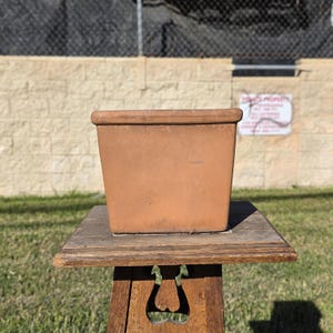 May include: A square terracotta planter with a smooth, matte finish and a slightly wider, flat rim. The pot features a uniform, earthy orange-brown color and a simple, clean-lined design, showcasing a natural clay texture throughout its exterior surface.