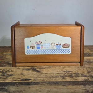 May include: A rectangular wooden bread box with a roll-top door. The front panel features a decorative white insert with a blue checkered border and a colorful graphic illustration of a strawberry, a basket, a heart, wheat, and a pie.