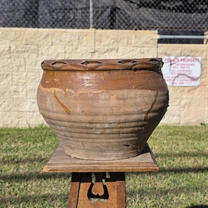 May include: A large, round, brown earthenware pot with a wide, wavy-edged rim. The exterior features a smooth, glazed upper section and a textured, ribbed lower body with visible signs of wear and earthy, weathered surface details.