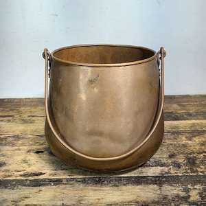 May include: A round copper pot with a smooth, metallic finish and a slightly weathered surface. The pot features a wide, open top and a sturdy, arched metal handle attached to two side loops, allowing for easy carrying and hanging display.