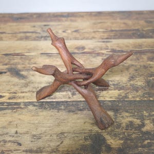 May include: A handcrafted wooden stand with a unique, intertwined design. The stand is made of dark brown wood and features three legs that converge at the center, creating a stable base. The stand is likely used for displaying decorative items.