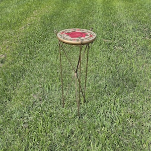 May include: A small, decorative plant stand with a round top featuring a red rose design. The stand has a gold-colored metal frame with three legs and a decorative cross-shaped support. The stand is set on a bed of green grass.