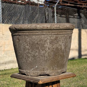 May include: A weathered, cylindrical brown clay planter with a thick, rounded rim and a textured, earthy surface. The pot features a wide opening, a tapered body, and a sturdy base with small feet, showing signs of age and natural wear throughout.