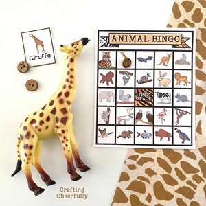 May include: A giraffe toy with brown spots on a yellow background. The giraffe is standing on a white surface. There is a bingo card with animal illustrations and the text "ANIMAL BINGO" on it. There are also two brown buttons on the surface.