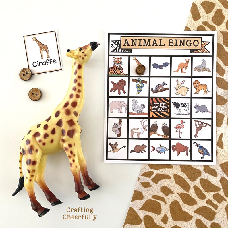 May include: A giraffe toy with brown spots on a yellow background. The giraffe is standing on a white surface. There is a bingo card with animal illustrations and the text "ANIMAL BINGO" on it. There are also two brown buttons on the surface.