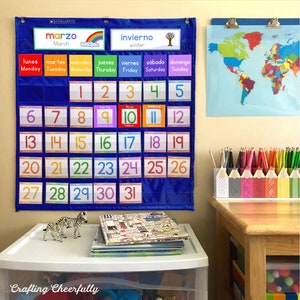 May include: A blue calendar with a rainbow border and the word "marzo" (March) in Spanish. The calendar has the days of the week in Spanish and English, and the numbers 1 through 31. There are two zebra figurines on a white shelf below the calendar.