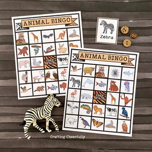 May include: Two printable animal bingo cards featuring a zebra, tiger, giraffe, elephant, flamingo, spider, and other animals. The cards are on a brown striped background with a zebra figurine and buttons.