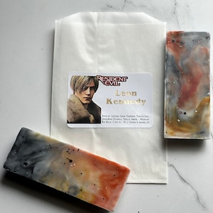 Puede incluir: Dos barras rectangulares de jab&oacute;n hecho a mano con un dise&ntilde;o marmoleado en tonos grises, naranja y marr&oacute;n. El jab&oacute;n est&aacute; etiquetado como "Resident Evil Leon Kennedy" con notas de cuero, cedro, teca, hoja de tabaco, s&aacute;ndalo, pachul&iacute;, vainilla, &aacute;mbar y cal&eacute;ndula. El jab&oacute;n pesa 3 oz y se vierte en el sur de California.